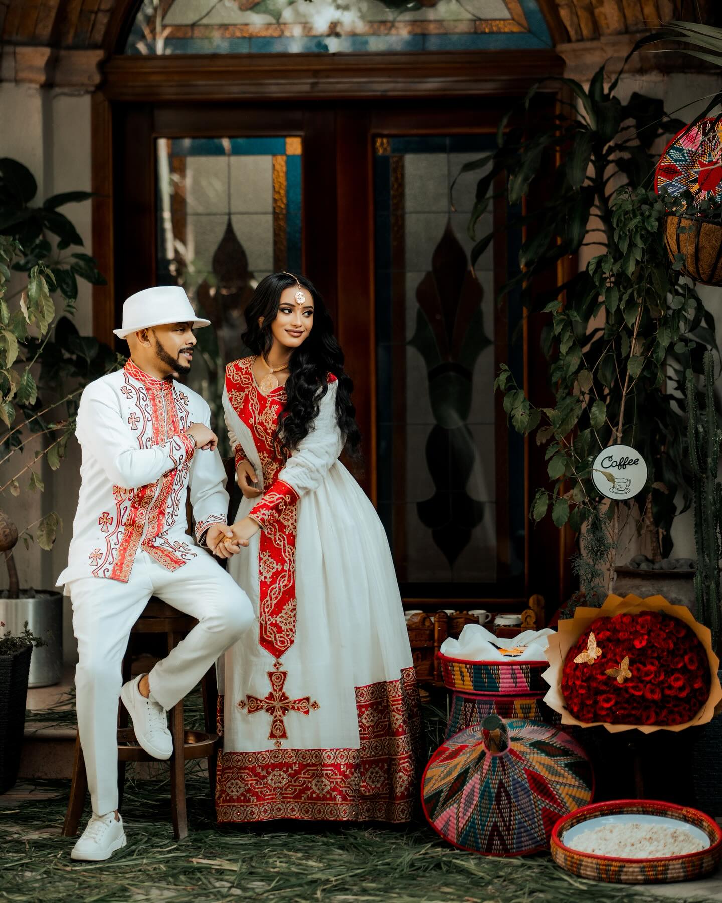 Couple in traditional attire standing in a decorative interior setting with plants and a stained glass window.