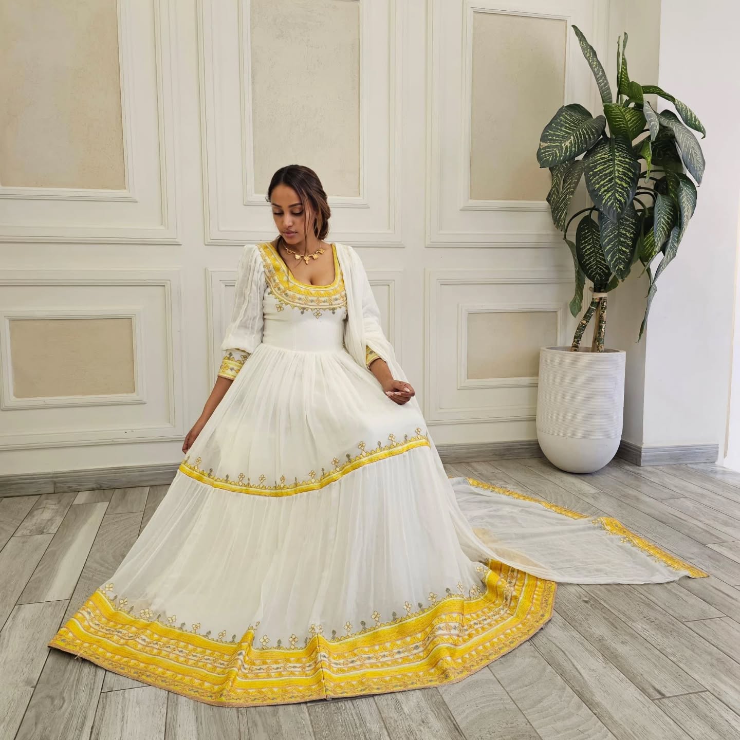 Woman in a white and yellow traditional outfit standing in a room with decorative walls and a plant.