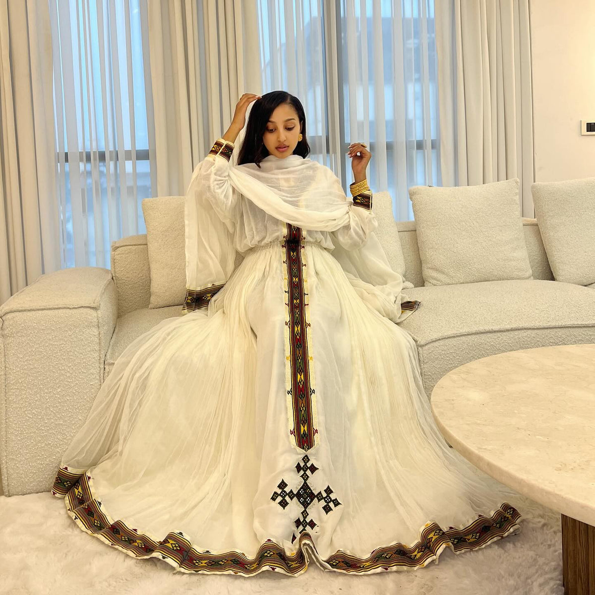 Woman in a long, elegant white dress with intricate patterns sitting on a couch in a modern living room.