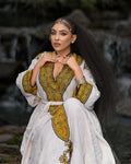 Woman wearing a white and gold traditional outfit with jewelry, standing against a natural background.