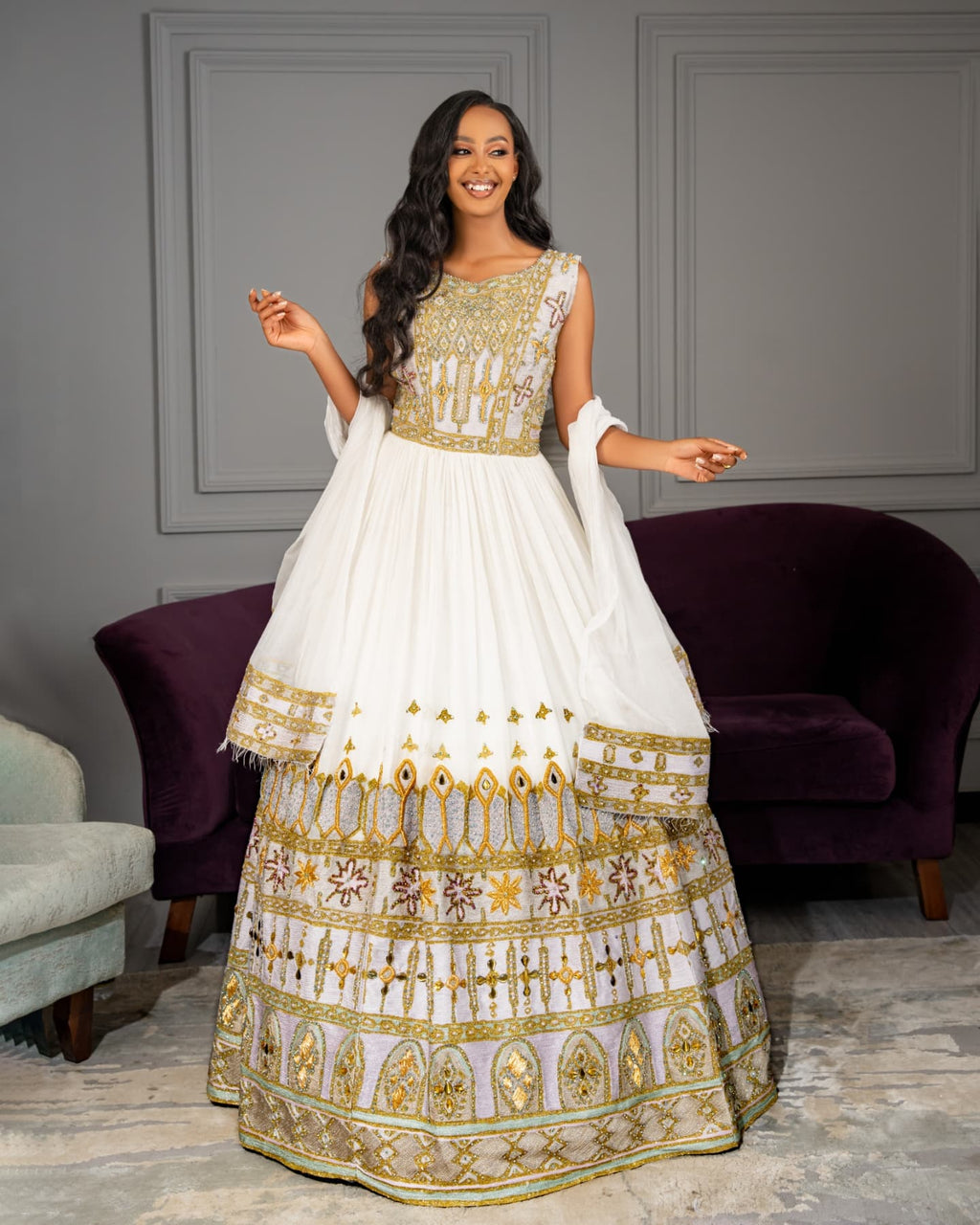 Woman wearing a white and gold traditional outfit in a room with purple chairs.