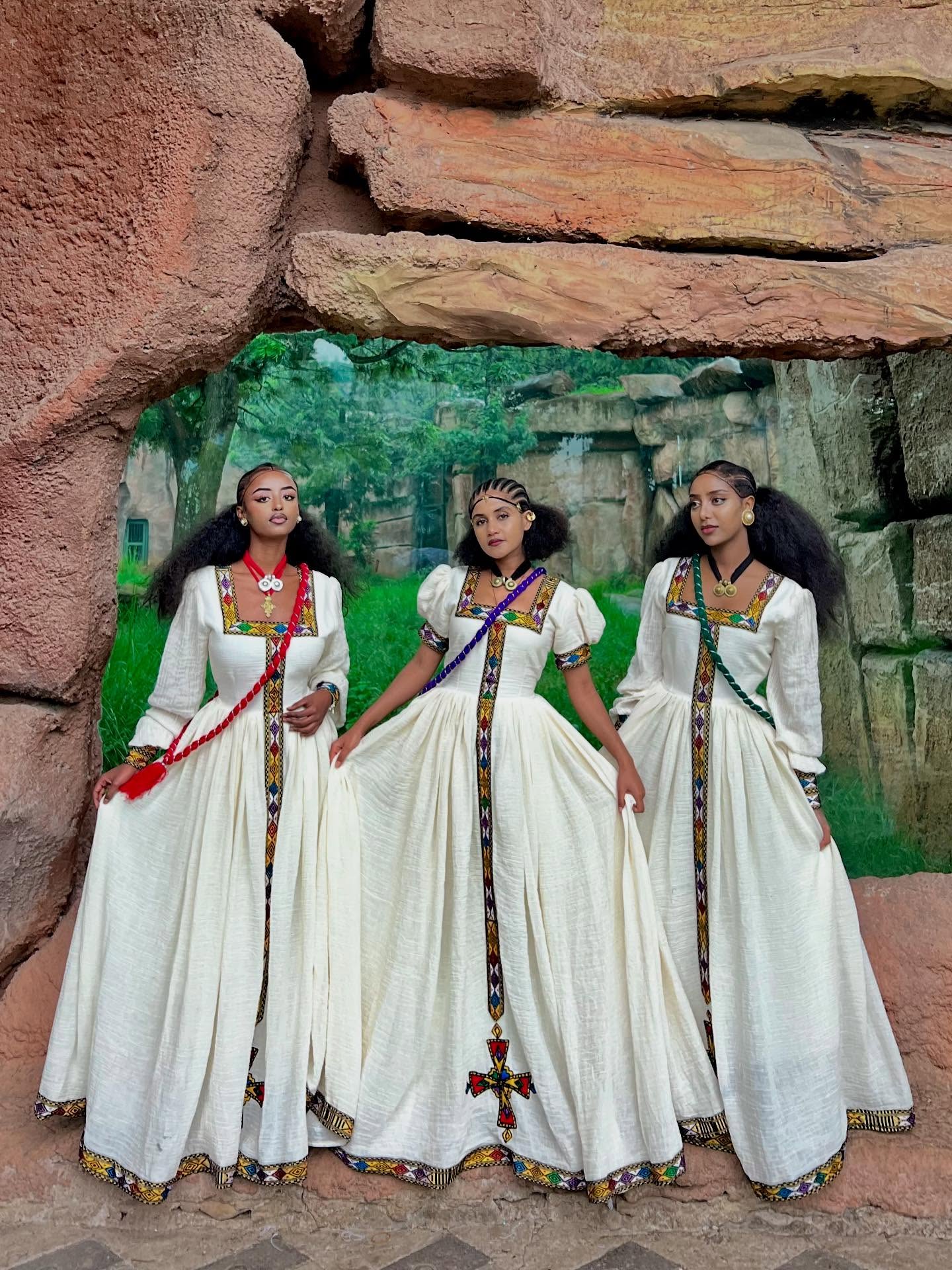 Three women in white traditional dresses with colorful accents standing in front of a stone wall.