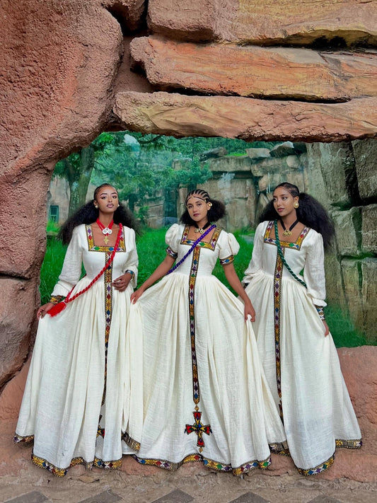 Three women in white traditional dresses with colorful accents standing in front of a stone wall.