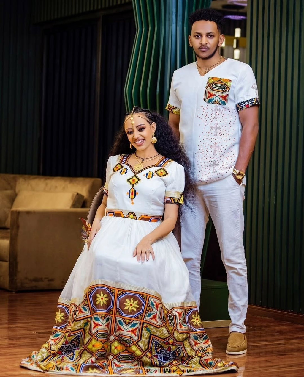 Two people in traditional attire standing in a room with a couch and decorative elements.