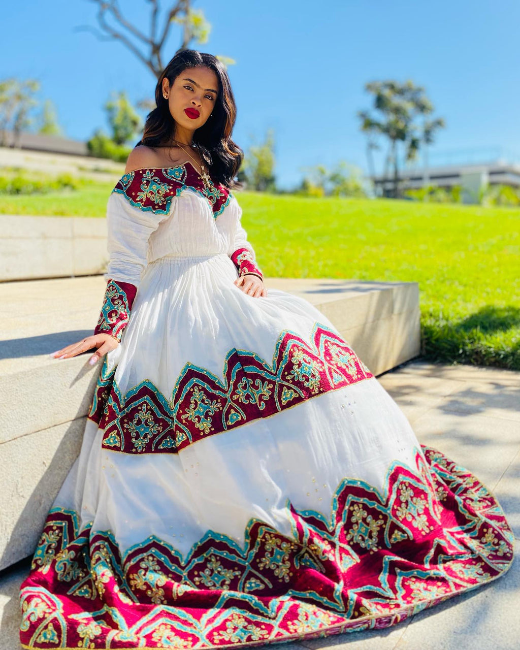 Woman in a white dress with colorful patterns sitting outdoors on a sunny day.