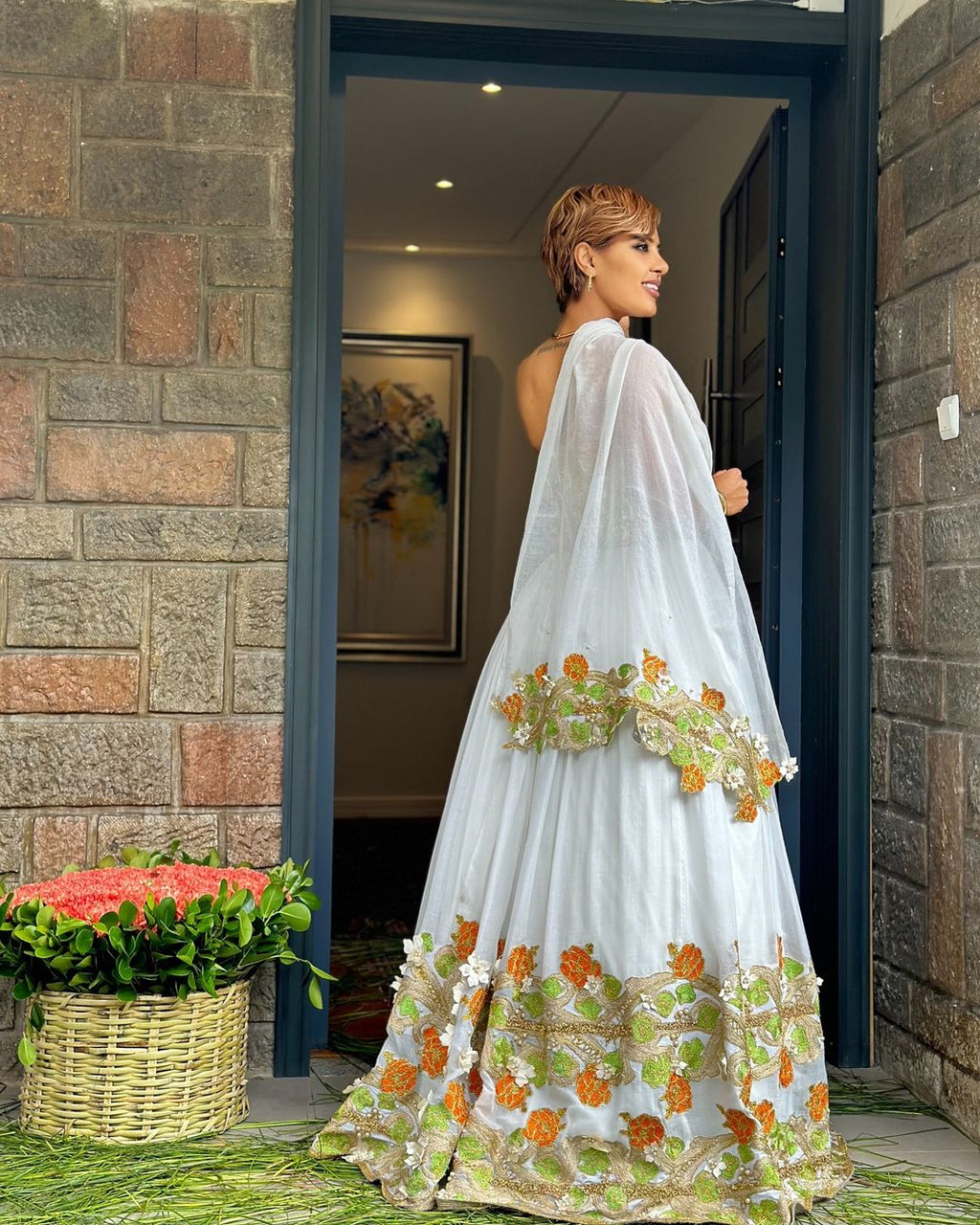 Woman in a white embroidered saree standing in a doorway with flowers in the foreground.