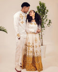 Couple in matching white outfits with gold patterns standing next to a plant.