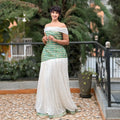 Woman in a green and white traditional outfit standing outdoors with plants in the background