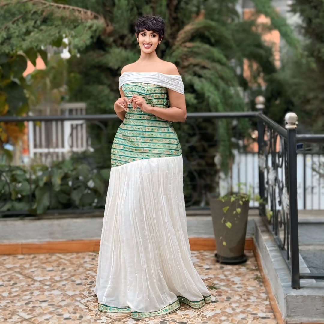 Woman in a green and white traditional outfit standing outdoors with plants in the background