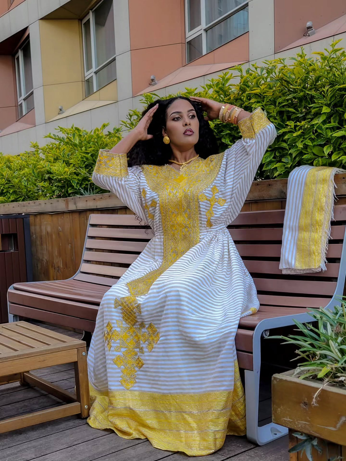 Woman in a yellow and white dress sitting on a bench with greenery and a building in the background