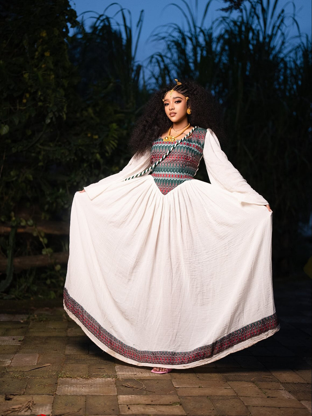 Woman in a traditional outfit with a long white skirt and patterned top standing outdoors.