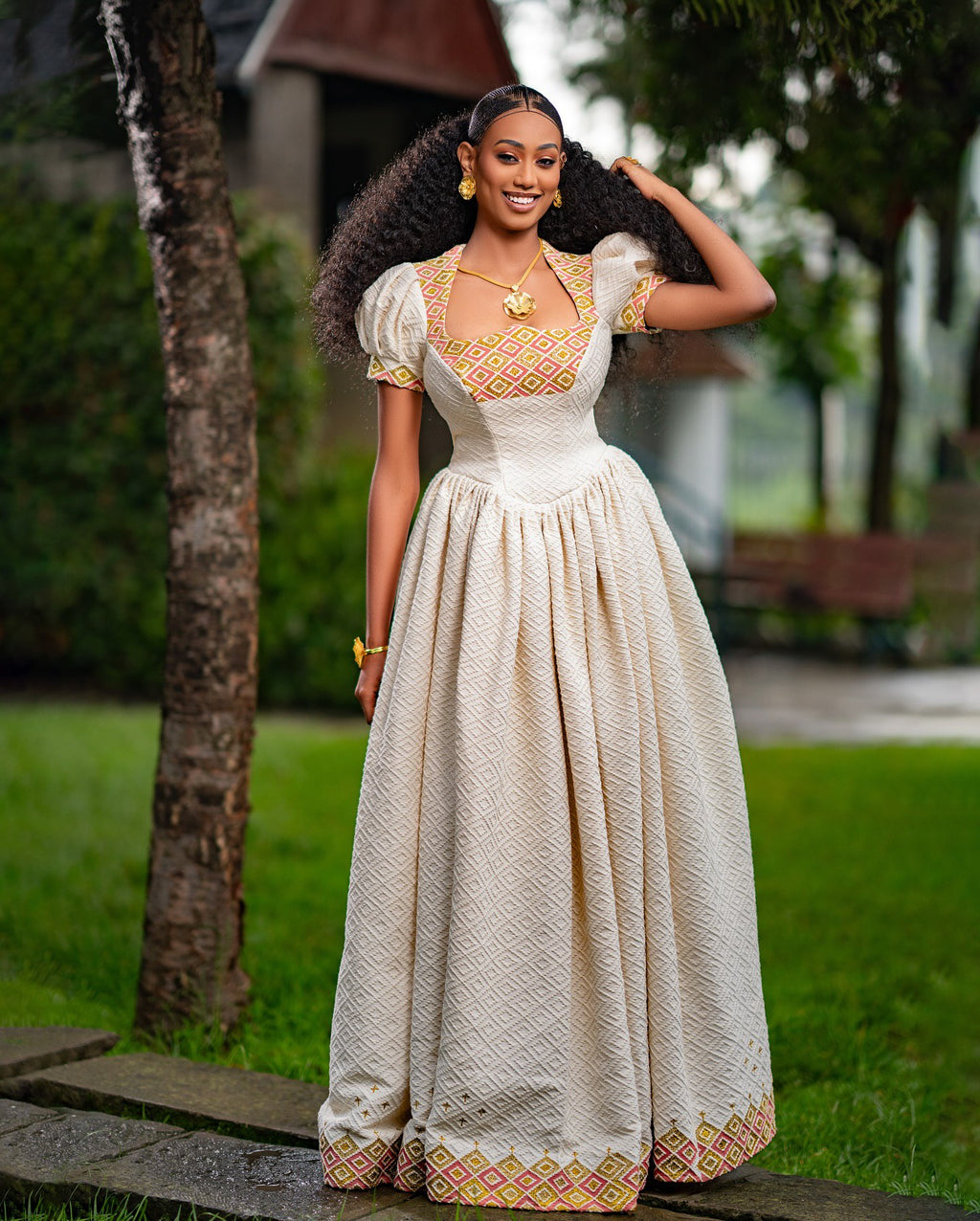 Woman in a traditional outfit standing outdoors with greenery in the background