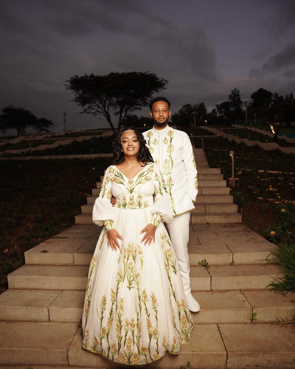 Couple in formal attire standing on stone steps outdoors at dusk.