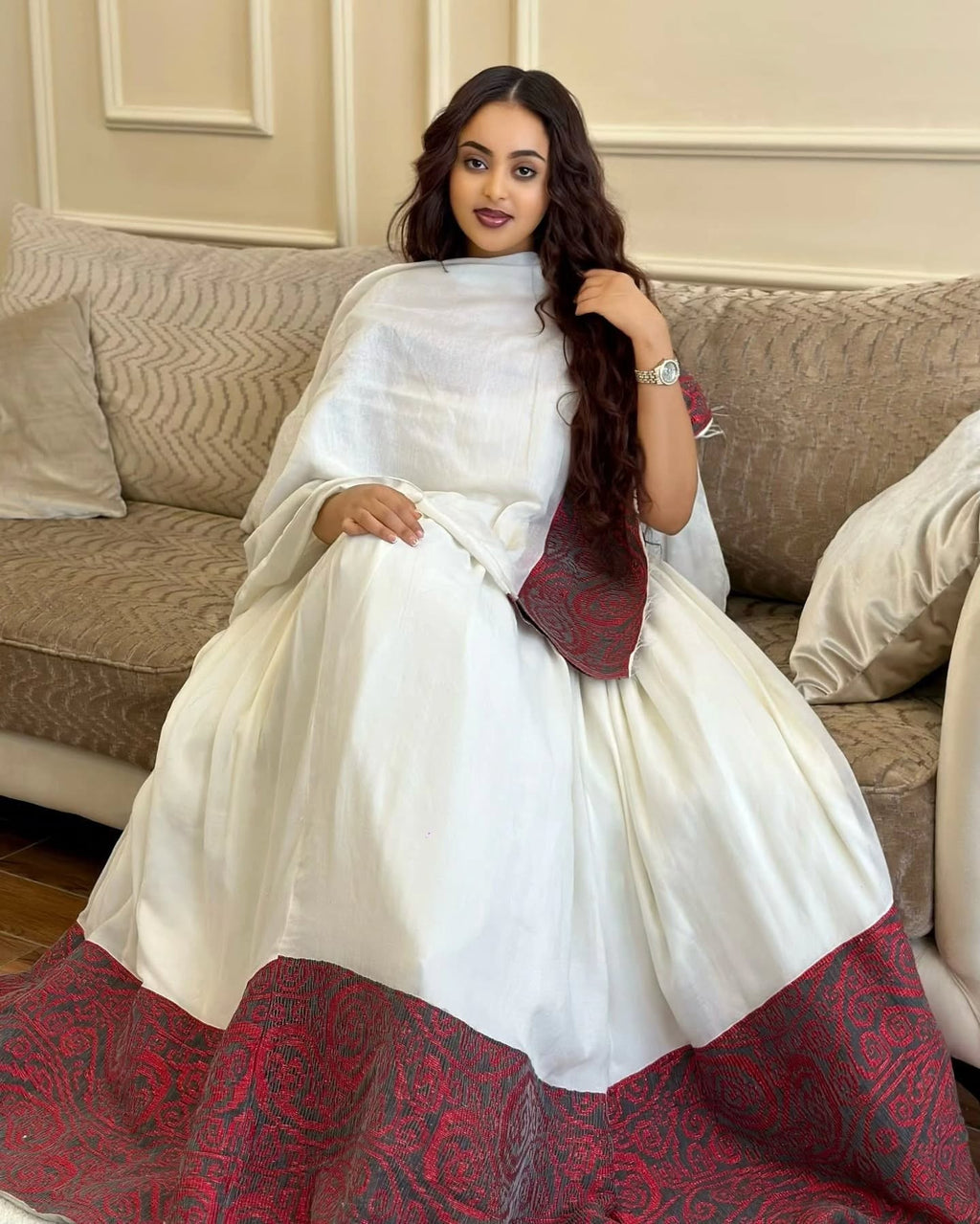 Woman in a white saree with red border sitting on a couch.