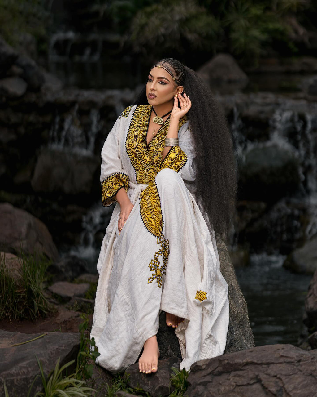 Woman in a white and gold traditional outfit standing by a rocky stream.