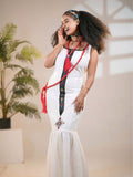 Woman in a white dress with decorative elements, standing indoors with a plant in the background.