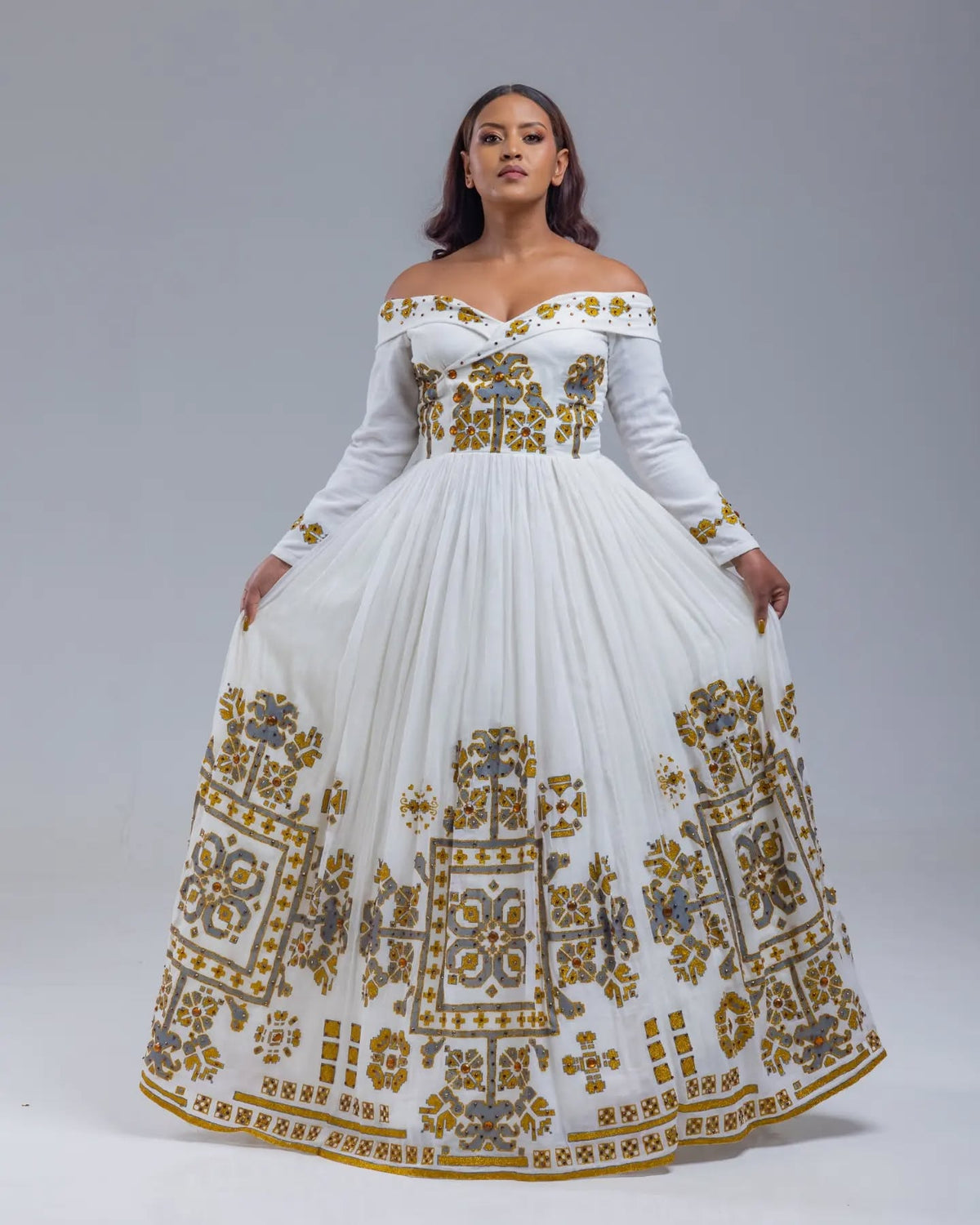 Woman wearing a white off-shoulder dress with gold embroidery on a gray background