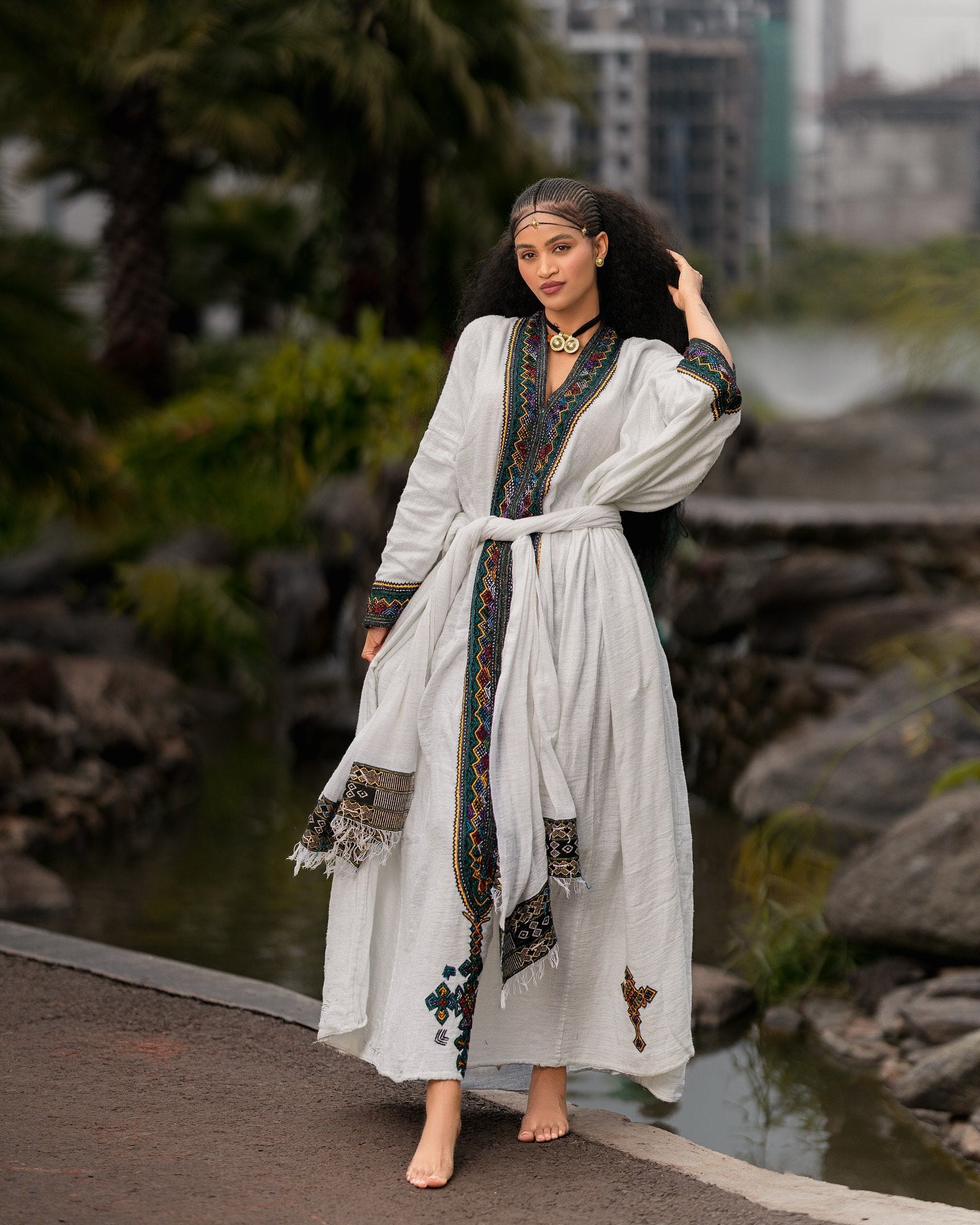 Woman in a long, white dress with decorative patterns standing by a body of water with rocks and greenery.