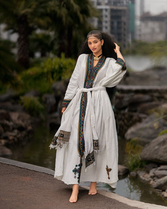 Woman in a long, white dress with decorative patterns standing by a body of water with rocks and greenery.